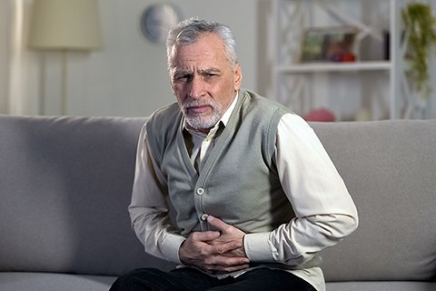 Älterer Mann mit Bauchschmerzen sitzt auf Sofa, hält sich den Bauch.