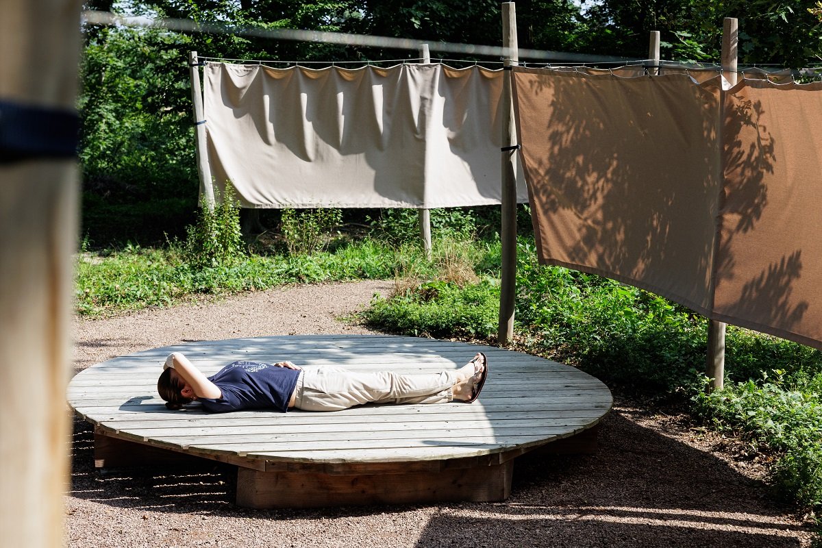 Person entspannt auf Holzplattform in besinnlichem Krankenhausgarten mit Stoffbahnen.