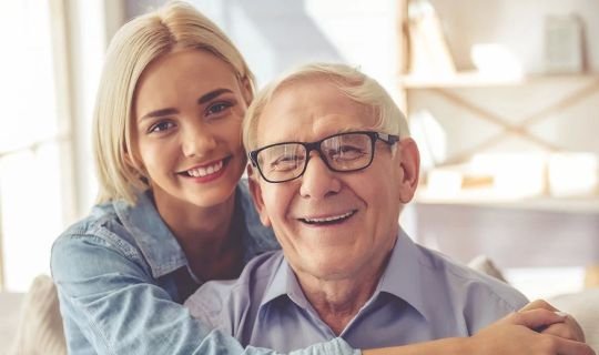 Junge Frau umarmt älteren Mann, beide lächeln in die Kamera.