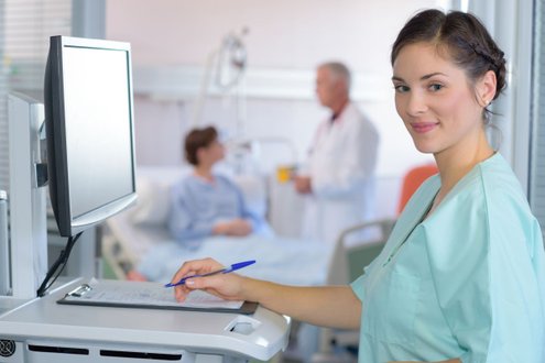 Pflegekraft in Krankenhaus, lächelnd, arbeitet mit Patientenakten am Computer.