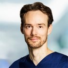 Porträt eines Mannes in blauer medizinischer Kleidung, neutraler Hintergrund.