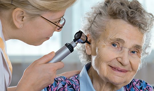 Ärztin untersucht Ohr einer älteren Patientin mit Otoskop.