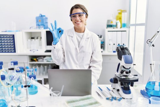 Lächelnde Forscherin im Labor zeigt 'OK'-Geste, umgeben von Laborausrüstung.
