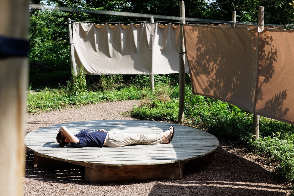 Person entspannt auf Holzplattform in besinnlichem Krankenhausgarten mit Stoffbahnen.