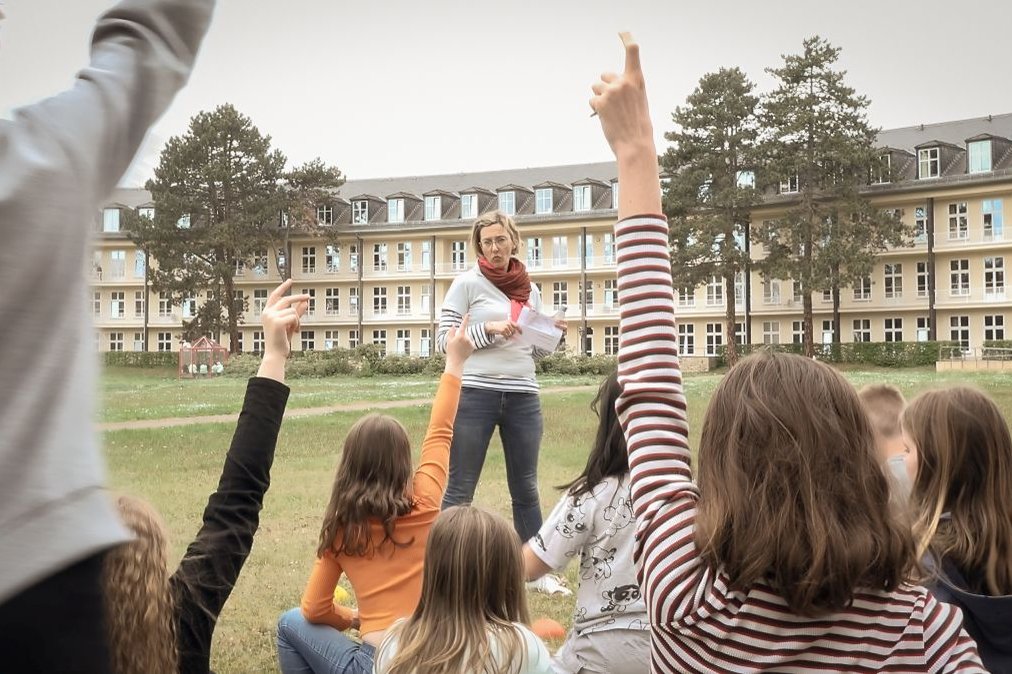 Kinder heben die Hand, während einer Lehrerin vor einem Krankenhaus zuhört.