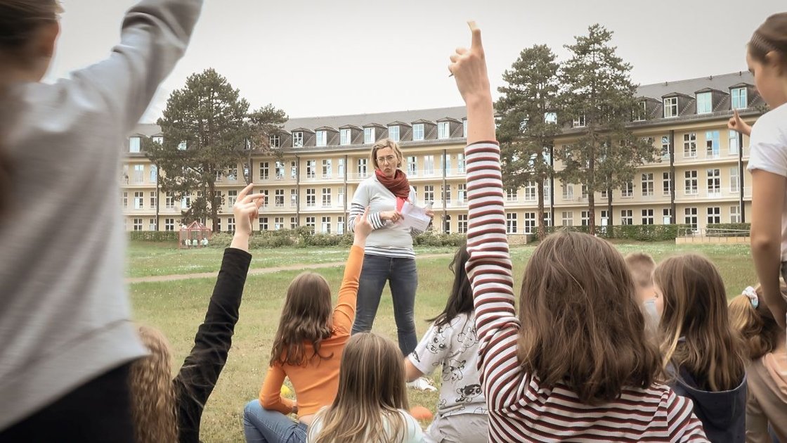 Kinder heben die Hand, während einer Lehrerin vor einem Krankenhaus zuhört.