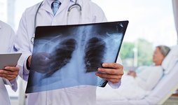 Arzt hält Röntgenbild, Patientin liegt im Krankenhausbett im Hintergrund.