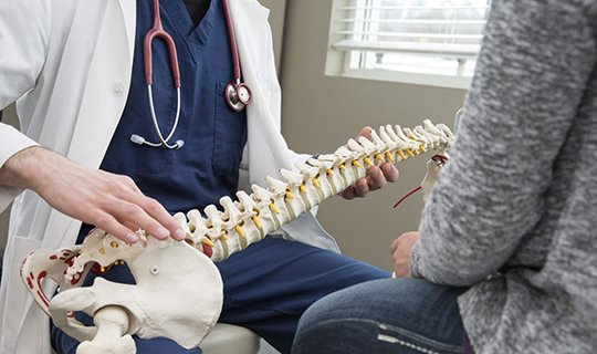 Arzt erklärt Wirbelsäulenmodell einem sitzenden Patienten in medizinischer Umgebung.