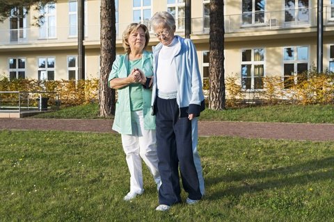 Pflegekraft unterstützt ältere Frau beim Spaziergang im Garten der Palliativstation.