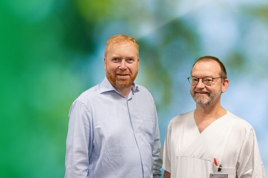 Zwei Männer vor grünem Hintergrund, lächelnd, eine trägt einen weißen Kittel.